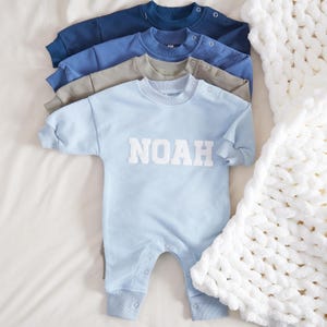 May include: A light blue baby onesie with the name "NOAH" in white letters. The onesie is folded and stacked on top of other onesies in blue and grey.