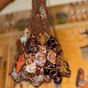 May include: A collection of raw gemstones, including amethyst, quartz, and obsidian, are wrapped in copper wire and suspended from copper chains. The gemstones vary in color, with shades of purple, orange, black, and white. The jewelry is displayed against a blurred background.