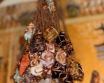 May include: A collection of raw gemstones, including amethyst, quartz, and obsidian, are wrapped in copper wire and suspended from copper chains. The gemstones vary in color, with shades of purple, orange, black, and white. The jewelry is displayed against a blurred background.