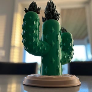 May include: A green cactus-shaped planter with two small green plants on top. The planter has a textured surface and a white base.