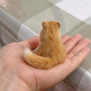 Pode incluir: Uma pequena figura de gato laranja com uma cauda e pelo fofo. O gato est&aacute; sentado, visto por tr&aacute;s, na palma de uma m&atilde;o. O pelo do gato &eacute; de cor laranja clara e a cauda tem uma ponta branca.