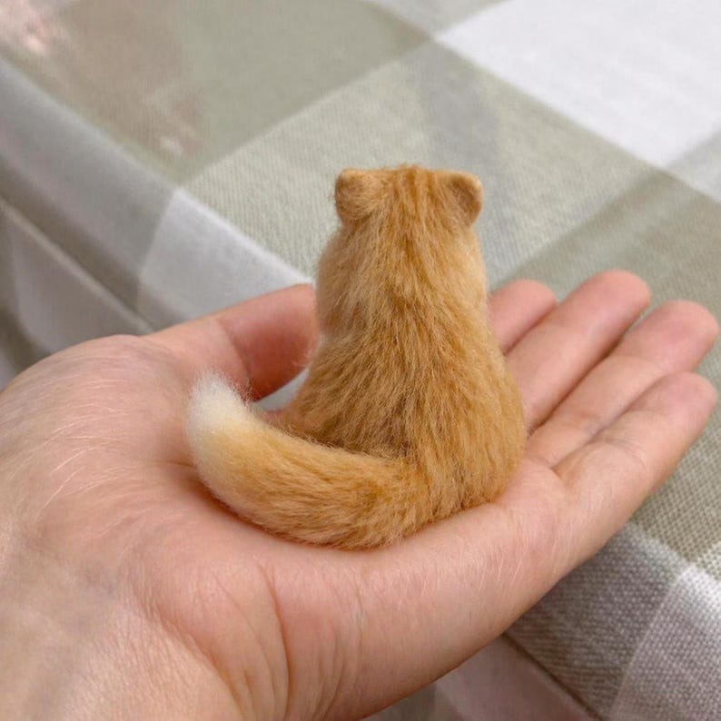 Pode incluir: Uma pequena figura de gato laranja com uma cauda e pelo fofo. O gato est&aacute; sentado, visto por tr&aacute;s, na palma de uma m&atilde;o. O pelo do gato &eacute; de cor laranja clara e a cauda tem uma ponta branca.