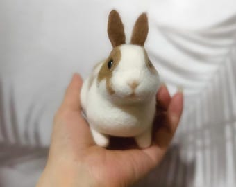 May include: A handmade felt bunny rabbit with brown and white markings. The bunny has upright brown ears and is held in a person's hand. The bunny is a cute, whimsical, and decorative item.