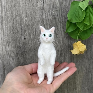 Può includere: Una piccola statuetta di gatto in lana infeltrita bianca, in piedi sulle zampe posteriori. Il gatto ha occhi verde brillante, un naso rosa e lunghi baffi bianchi. Il pelo ha un aspetto morbido e testurizzato, con una coda lunga e sottile.