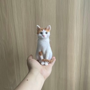 Peut inclure: Figurine de chat blanc et orange fait à la main. Le chat est assis, la queue enroulée autour de ses pattes. Il présente des marques orange sur les oreilles, la tête et la queue. La figurine est tenue dans une main devant un fond en bois brun clair.