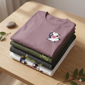 May include: A stack of four folded t-shirts in various colors: mauve, olive green, charcoal gray, and white. Each shirt features a cartoon graphic on the front. The top shirt has a cow graphic, while the others have different cartoon characters.
