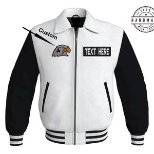 Puede incluir: Chaqueta de béisbol blanca y negra con un parche de águila gris en el lado izquierdo y un parche negro con el texto "TEXT HERE" en el lado derecho. La chaqueta tiene una cremallera y un cuello, puños y dobladillo de canalé.