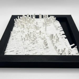 May include: A 3D-printed white cityscape model, framed in a black square. The model depicts a detailed urban landscape with numerous buildings and structures. The frame adds a clean, modern aesthetic to the artwork.