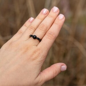 Puede incluir: Un anillo negro con una cuenta central redonda y una banda geométrica. El anillo se muestra en una mano, con un fondo natural borroso. El diseño del anillo es simple y elegante, adecuado para el uso diario.