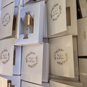 Op de afbeelding: Witte cadeauboxen met een gouden sticker met de initialen "M&L" en de datum "02-20-2025". De dozen bevatten kleine, heldere glazen flesjes met gouden dopjes.