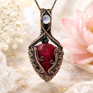 May include: A pendant featuring a large, faceted, deep red ruby-coloured stone encased in intricate, dark copper wire wrapping. A small, round, iridescent moonstone is set above the main stone. The pendant hangs from a matching copper chain.