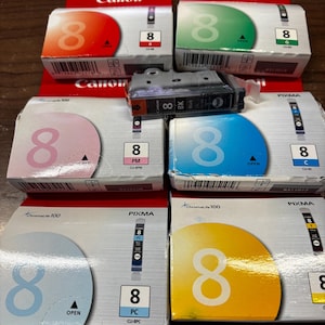 May include: A collection of Canon Pixma 8 ink cartridges in various colors including red, green, photo magenta, cyan, photo cyan, and yellow, with one black ink cartridge resting on top of the boxes, all featuring the number 8 and ChromaLife 100 text.