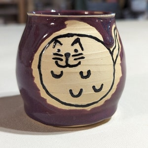 May include: A purple ceramic mug with a hand-painted design of a cat with a white face and black eyes. The cat is outlined in black and has a white tail.