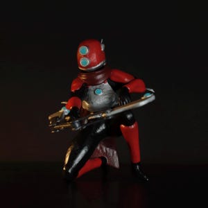 May include: A red and black figurine of a futuristic character in a kneeling pose, holding a silver weapon. The character has a red helmet with two blue eye-like features and a silver chest plate. The figure is set against a dark background.