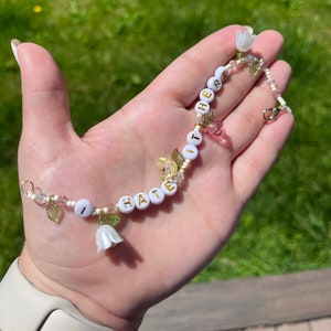 Puede incluir: Un brazalete de cuentas blancas con las palabras "I HATE IT HERE" escritas en letras doradas. El brazalete está decorado con pequeñas flores blancas y hojas verdes.