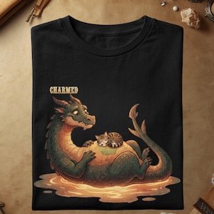 Puede incluir: Una camiseta negra con una ilustración gráfica de un dragón verde tumbado en un charco de luz brillante. Un pequeño gato atigrado marrón duerme acurrucado sobre la barriga del dragón. La palabra "CHARMED" está impresa en color dorado arriba.
