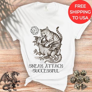 Può includere: T-shirt bianca con un'illustrazione in bianco e nero di un gatto che attacca un drago e un dado D20. Il testo "SNEAK ATTACK SUCCESSFUL" è sotto la grafica. Un cerchio rosso con "FREE SHIPPING TO USA" è in alto a destra.