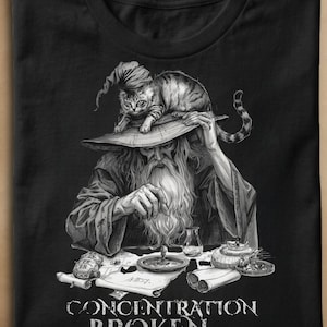Peut inclure: Un t-shirt noir avec un graphique monochrome détaillé représentant un magicien barbu portant un chapeau pointu sur lequel est perché un chat tigré. Le magicien est assis à une table avec des parchemins et des potions, texte CONCENTRATION BROKEN.