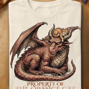 Peut inclure: Un t-shirt couleur crème arborant une illustration détaillée d'un grand dragon brun aux écailles, avec un petit chat tigré orange reposant entre ses cornes. Sous l'illustration, le texte indique : PROPERTY OF THE ORANGE CAT.