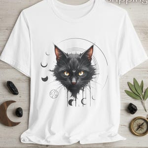 Puede incluir: Camiseta blanca con una ilustración detallada de un gato negro con ojos amarillos y un tercer ojo, rodeado de diseños celestiales. El pelaje del gato está intrincadamente representado, y el diseño incluye lunas crecientes y un patrón circular.