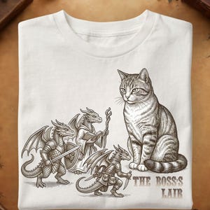 May include: A white t-shirt featuring a detailed brown ink illustration of a large tabby cat sitting next to three small, armored dragon-like creatures. The text "THE BOSS'S LAIR" is printed in a brown serif font near the bottom right corner.