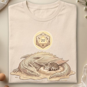 Peut inclure: Un t-shirt beige avec une impression graphique représentant un dragon endormi enroulé autour d'un petit chat tigré reposant sur un coussin. Au-dessus du dragon, un dé à vingt faces est illustré avec le chiffre 20 centré dans un cercle jaune lumineux.