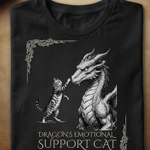 Peut inclure: Un t-shirt noir avec une illustration au trait blanc représentant un chat tigré tendant la patte vers le museau d'un grand dragon. Sous le dessin, le texte indique DRAGON'S EMOTIONAL SUPPORT CAT. Le motif inclut des ornements aux coins.