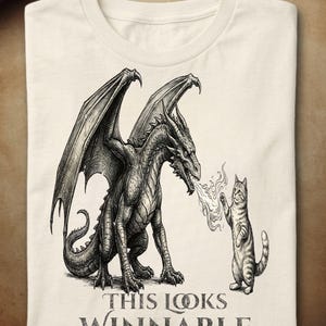 Peut inclure: Un t-shirt couleur crème arborant une illustration à l'encre noire et blanche représentant un dragon crachant du feu sur un chat tigré debout. Sous le dessin, le texte "THIS LOOKS WINNABLE" apparaît avec les deux "O" entrelacés en anneaux.