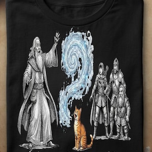 Könnte beinhalten: Ein schwarzes T-Shirt mit einer grafischen Illustration eines Zauberers, der einen blauen, wirbelnden Zauberspruch auf eine orangefarbene getigerte Katze richtet. Neben der Katze stehen ein Krieger, ein Ritter und ein Zwerg. Unten steht "WHO CAST THAT?".