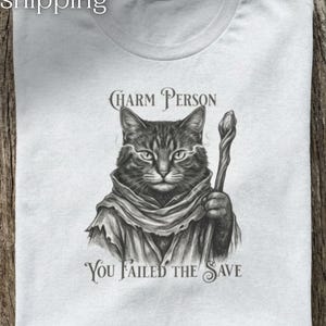 Könnte beinhalten: Hellgraues T-Shirt mit einer detaillierten Illustration einer Katze in einem Gewand, die einen Stab hält. Der Text "Charm Person" steht über der Katze, und "You Failed the Save" steht darunter.