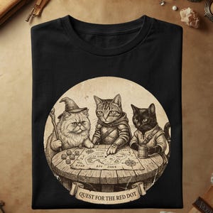 Może przedstawiać: Czarny t-shirt z okrągłą grafiką przedstawiającą trzy koty siedzące przy drewnianym stole z mapą i kośćmi. Koty są ubrane w stroje fantasy, a na banerze u dołu znajduje się wyraźny napis "QUEST FOR THE RED DOT".