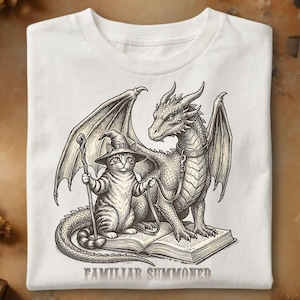 Peut inclure: Un t-shirt blanc avec une illustration en noir et blanc représentant un chat portant un chapeau de magicien et tenant un bâton, debout à côté d'un dragon sur un livre ouvert. Le texte FAMILIAR SUMMONED est imprimé sous l'illustration.