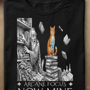 Peut inclure: Un t-shirt noir avec une illustration graphique représentant un magicien barbu attrapant des livres sur une étagère, tandis qu'un chat orange assis sur une pile de livres tient un cristal bleu. Le texte en bas indique ARCANE FOCUS NOW MINE.