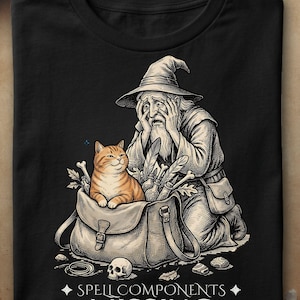 Puede incluir: Una camiseta negra con una ilustración gráfica de un mago con las manos en la cara, arrodillado junto a un bolso que contiene un gato atigrado naranja, plumas y huesos. Debajo de la imagen, el texto dice SPELL COMPONENTS MISSING.