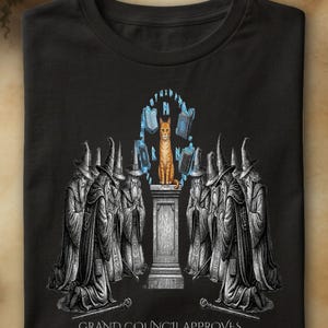 Peut inclure: Un t-shirt noir avec un motif représentant un chat orange assis sur un piédestal en pierre, entouré de livres flottants et de runes bleues lumineuses. En dessous, des personnages en robe et chapeau pointu sont agenouillés. Texte : GRAND COUNCIL APPROVES THE ORANGE ONE IS CHOSEN.
