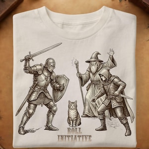 Può includere: Una t-shirt color crema con un'illustrazione a inchiostro marrone scuro di un cavaliere con spada e scudo, un mago che tiene un bastone e un ladro con un pugnale attorno a un gatto. Il testo "ROLL INITIATIVE" è stampato sotto il gatto.