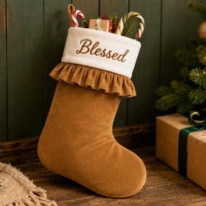 May include: A tan Christmas stocking featuring a white plush cuff embroidered with the word Blessed in brown script. The stocking has a ruffled trim below the cuff and is filled with candy canes and a small gift box wrapped in brown paper.