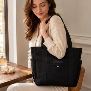 Op de afbeelding: Een zwarte canvas tas met twee stevige schouderbanden, zijvakken en een voorvak met een goudkleurige drukknoopsluiting. De naam Olivia is met donker garen op het voorvak van de tas geborduurd.