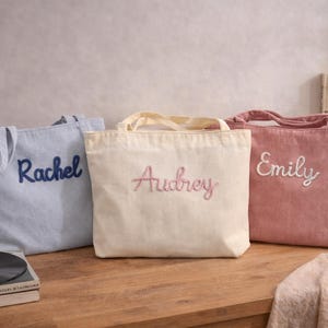 Puede incluir: Tres bolsas de mano personalizadas en azul, crema y rosa. Cada bolsa tiene un nombre bordado en un color complementario: "Rachel" en azul, "Audrey" en rosa y "Emily" en blanco. Las bolsas están sobre una superficie de madera.