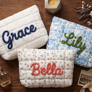 Puede incluir: Tres neceseres acolchados con nombres bordados. Uno es blanco con "Grace" en azul marino, otro es floral azul con "Lily" en verde, y el tercero es blanco con estampado de cerezas rojas y "Bella" en coral.