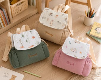 Op de afbeelding: Drie rugzakken van corduroy in olijfgroen, beige en oudroze. Elke tas heeft een klep met een uniek printpatroon en een geborduurde naam: Forest, Honey of Blush. De tassen zijn voorzien van verstelbare schouderbanden en zijvakken.