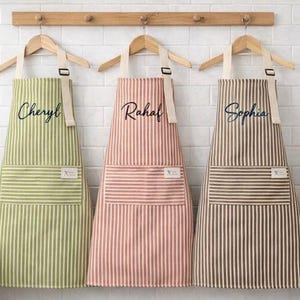 May include: Three personalized aprons with vertical stripes in green, pink, and brown, each with a name embroidered on the front. The aprons hang on wooden hangers against a white brick wall, ideal for cooking or baking.