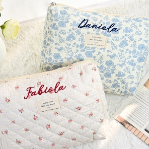 May include: Two quilted cosmetic bags. One is white with red floral embroidery and the name "Fabiola" in red. The other is light blue with a floral pattern and the name "Daniela" in blue. Both have a zipper closure.
