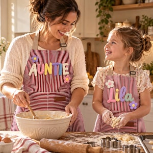 Puede incluir: Dos delantales a juego de rayas rojas y blancas con letras 3D personalizables. Un delantal muestra la palabra AUNTIE con una flor morada, y el otro muestra el nombre Lila con una flor blanca y un detalle de flor morada.