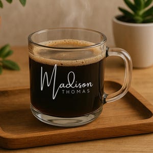 May include: A clear glass coffee mug filled with dark coffee, with the name "Madison Thomas" in white script. The mug sits on a wooden tray, with plants in the background.