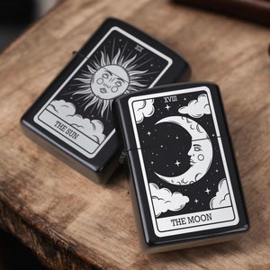 May include: Two black lighters with silver designs. The first lighter features a sun with the text "THE SUN" and the Roman numeral "XXI". The second lighter features a crescent moon with the text "THE MOON" and the Roman numeral "XVIII".