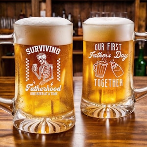 May include: Two clear glass beer mugs filled with beer and foam, sitting on a wooden bar. One mug reads "Surviving Fatherhood" with a skeleton graphic. The other mug says "Our First Father's Day Together".
