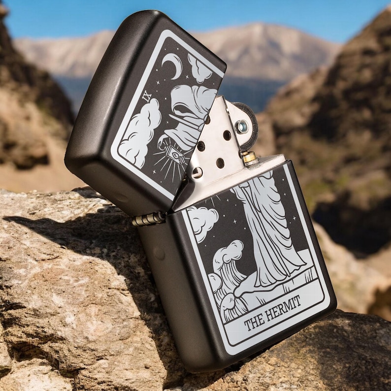 May include: Black lighter with a silver design of the Hermit tarot card. The card features a hooded figure holding a lantern and walking on a path with a crescent moon in the sky. The text "THE HERMIT" is printed at the bottom of the card.