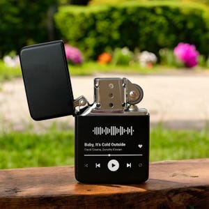 May include: Black lighter with a music player design. The lighter has a silver top and a black body with a white graphic of a music player interface. The screen displays the song "Baby, It's Cold Outside" by Frank Sinatra and Dorothy Kirsten.