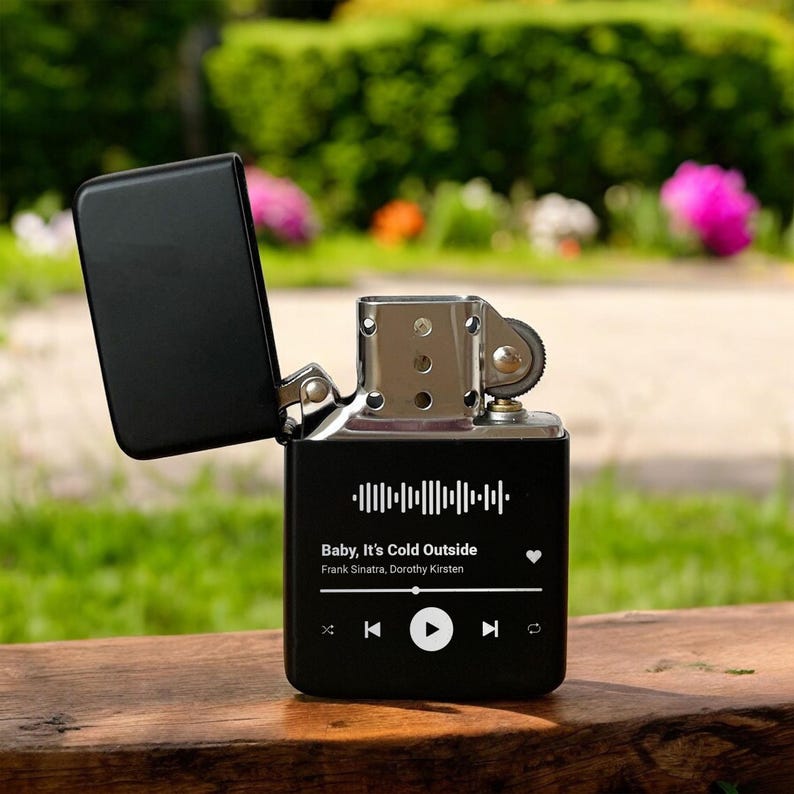 May include: Black lighter with a music player design. The lighter has a silver top and a black body with a white graphic of a music player interface. The screen displays the song "Baby, It's Cold Outside" by Frank Sinatra and Dorothy Kirsten.